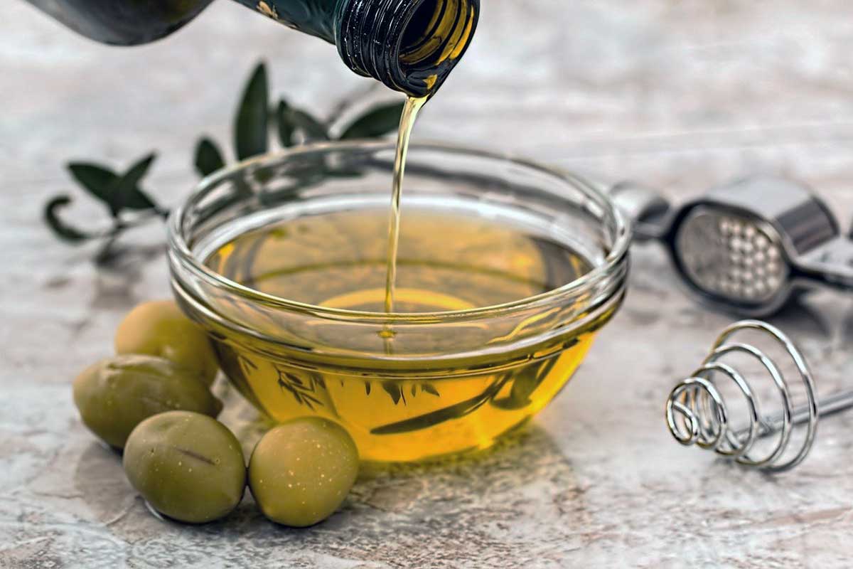 Olive oil being poured into glass bowl, from an olive oil bottle, surrounded by olives and kitchen tools on kitchen counter.
